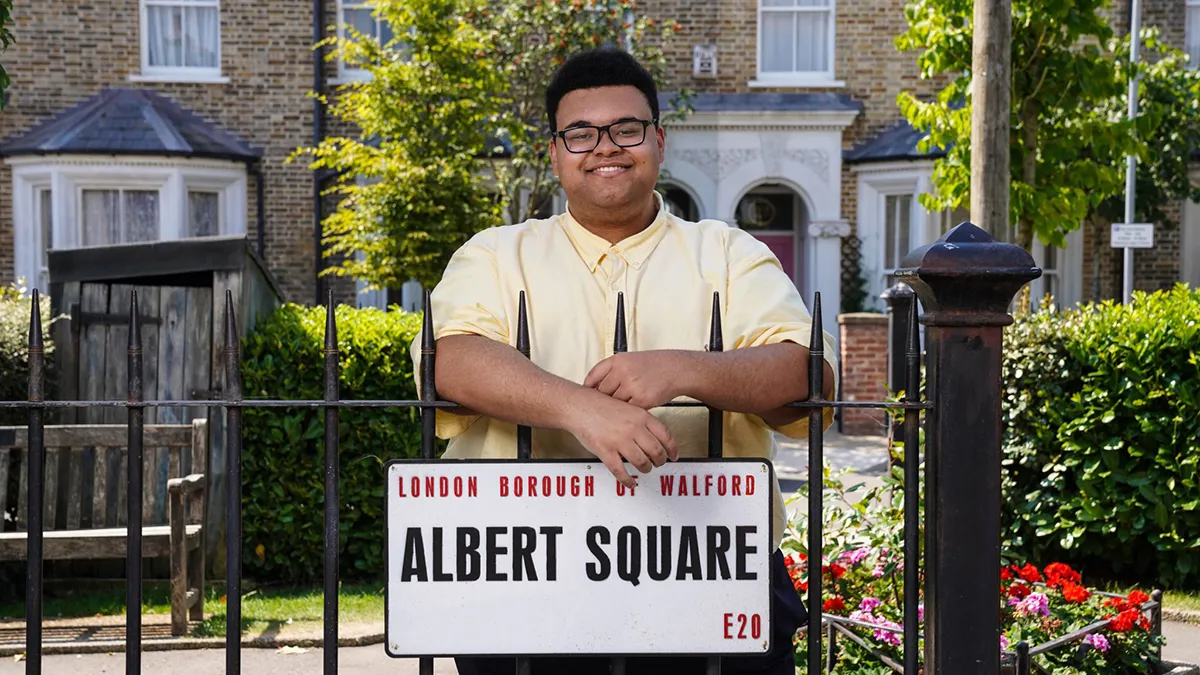 A Leeds Trinity University alumnus wearing a pale yellow shirt and smiling and posing behind a low iron fence with a white sign reading Albert Square on it..