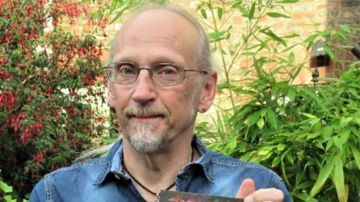 A man wearing glasses and blue shirt smiles while stood in front of green bushes.