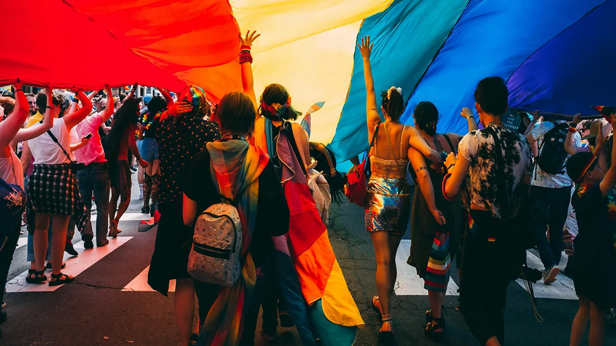 A group of people with their back turned to the camera, underneath a big rainbow flag and holding or wearing rainbow attire..
