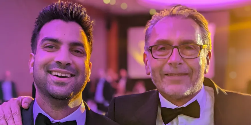 Two males in black tie suits stand next to each other smiling.