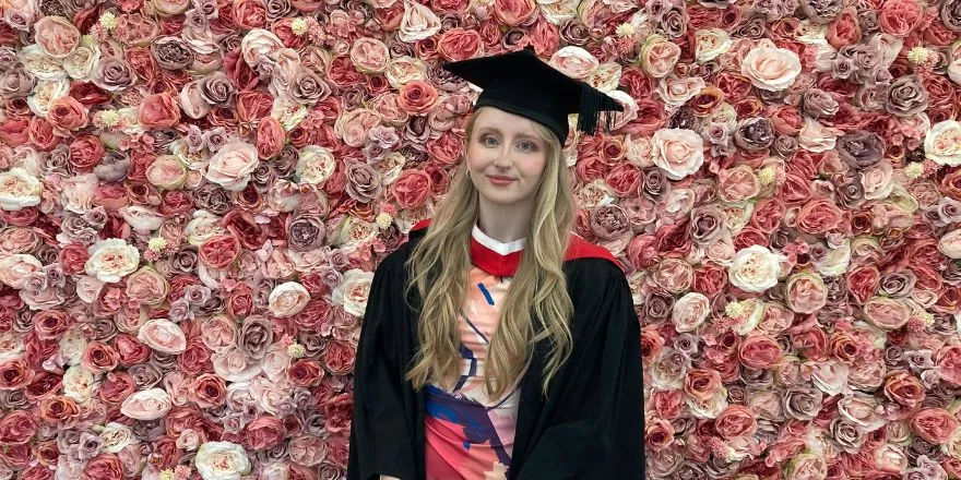 A girl with blonde hair wearing a black graduation cap and gown stands in front of wall of pink flowers.