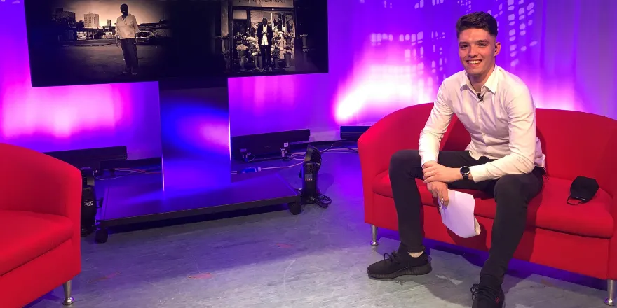 Male with brown hair, white shirt and dark trousers sits on red sofa to the right of tv screen.