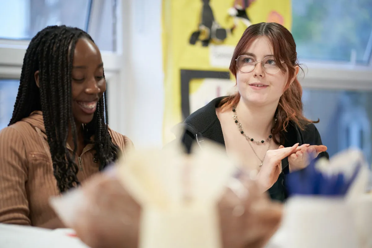Two education students sit together in a classroom, smiling and taking part in a group discussion..