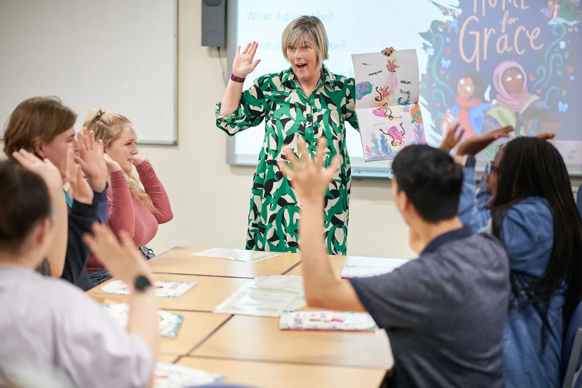 A lecturer enthusiastically reads a children’s book to education students, who raise their hands to join in the activity..