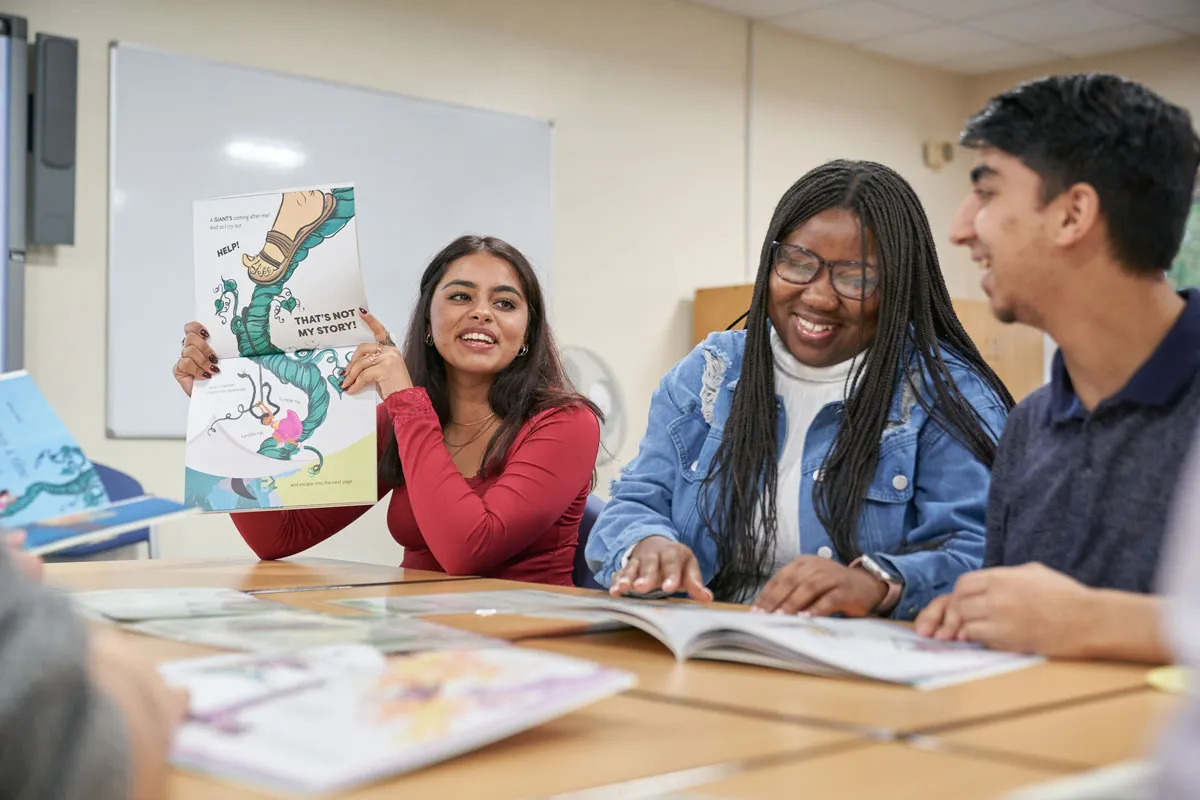 A group of students sit around a classroom table, holding and reading children’s picture books while smiling and discussing the content..