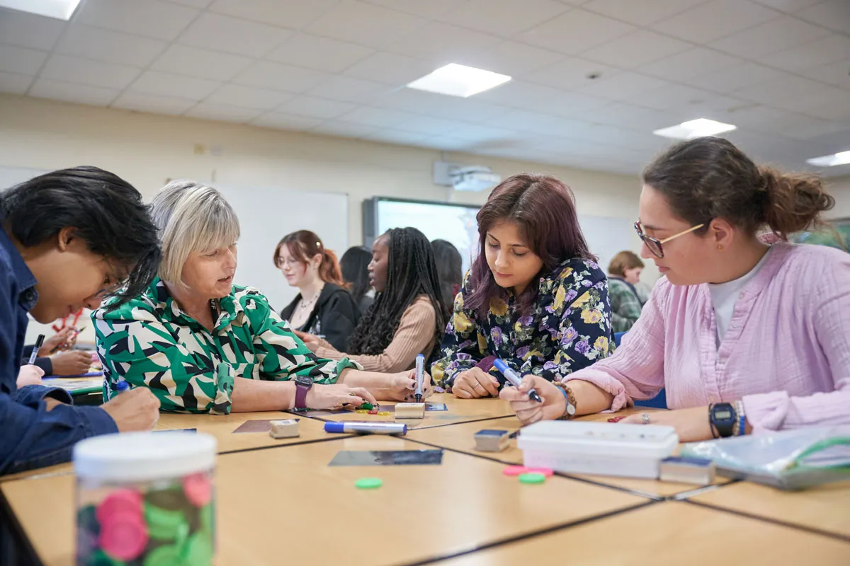 A lecturer and students work closely together at a classroom table, using pens, counters, and other teaching resources for an interactive activity..