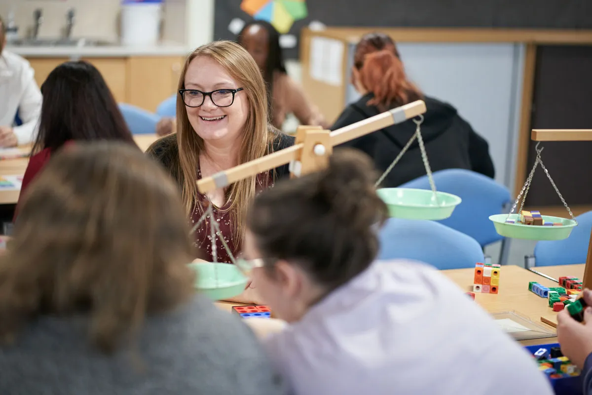 A student smiles while working with classroom resources, including scales and colourful counting blocks, during a group activity..