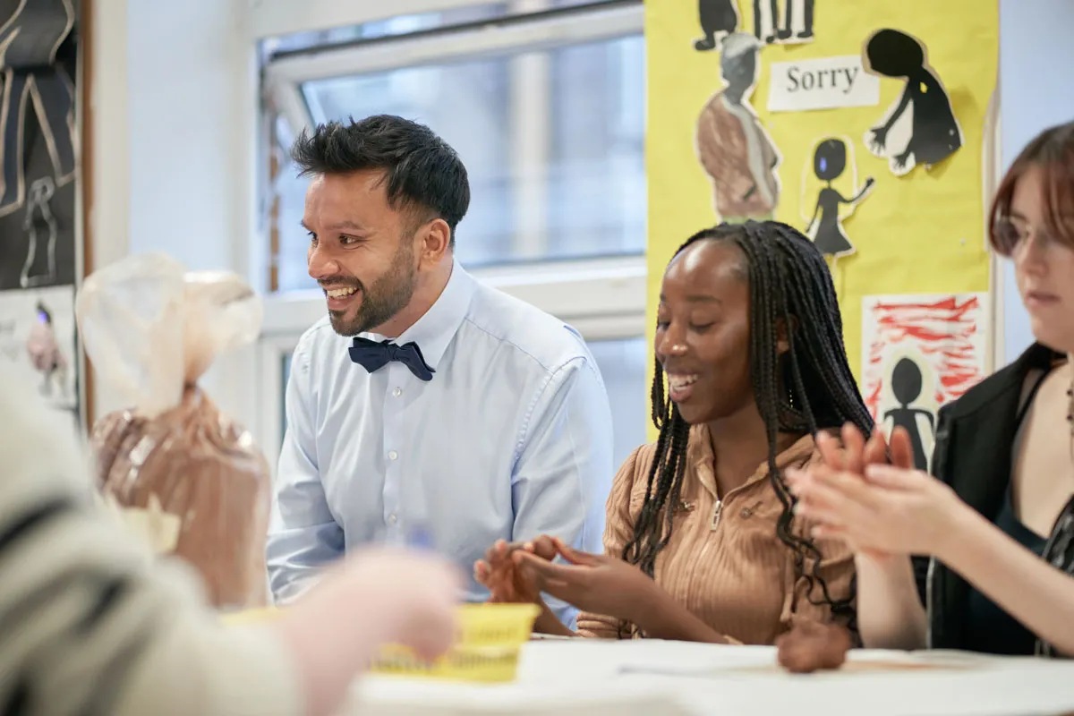 A lecturer and students sit at a table in a classroom, smiling and shaping clay during a practical activity. Colourful posters are visible on the wall in the background..