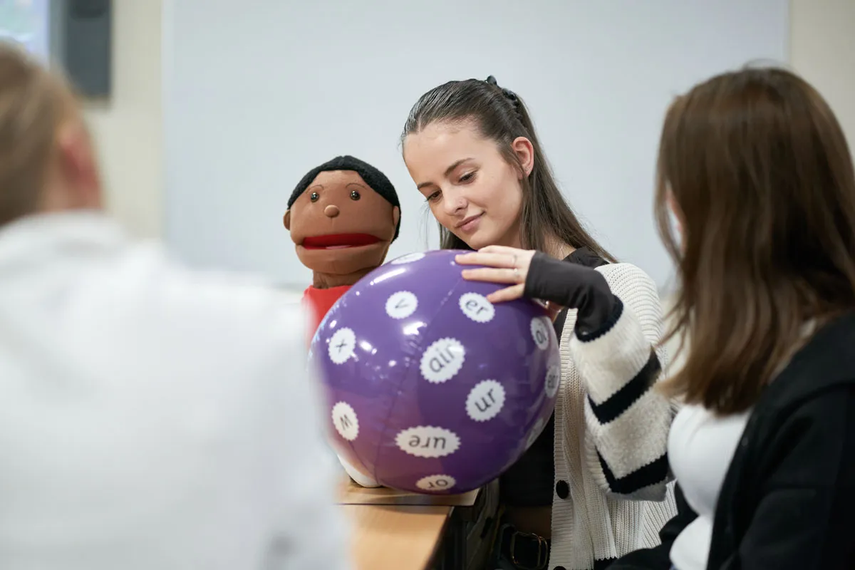 A student in a classroom holds a puppet and a large purple inflatable ball covered in phonics sounds like “ur,” “er,” and “oi,” while engaging in an activity with classmates..
