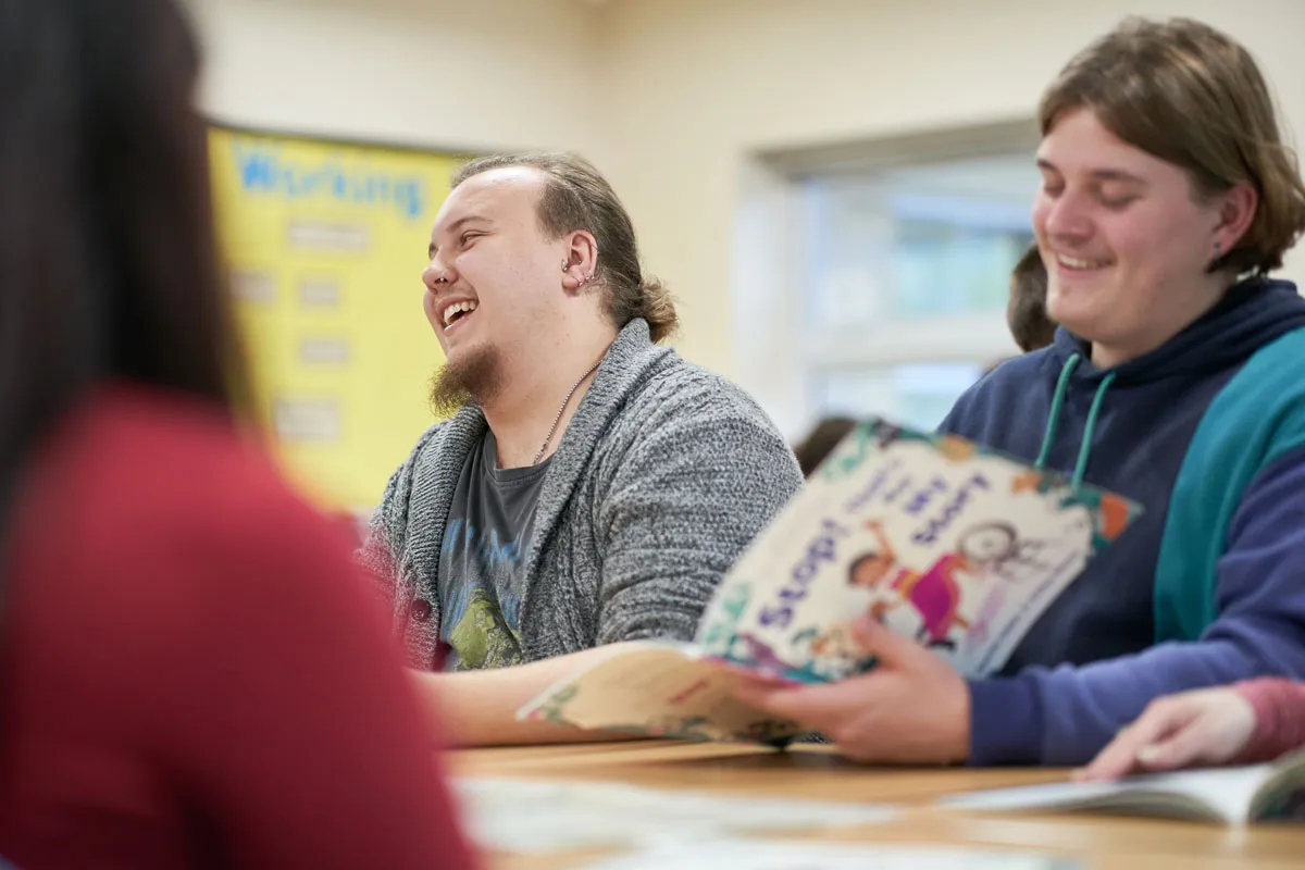 Two students sit at a classroom table, laughing and smiling while holding children’s storybooks during a lesson..