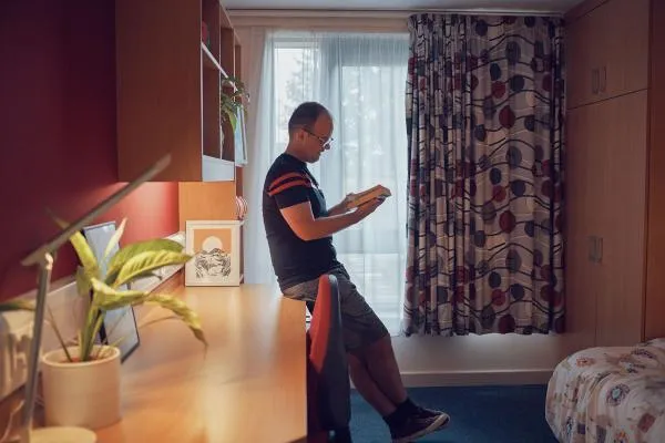 A student leans against a desk in a university accommodation room, reading a book. The room features a long wooden desk with a lamp, plant, and framed artwork, as well as shelving, a wardrobe, and a bed with a patterned duvet. Light filters in through large, patterned curtains covering a window..