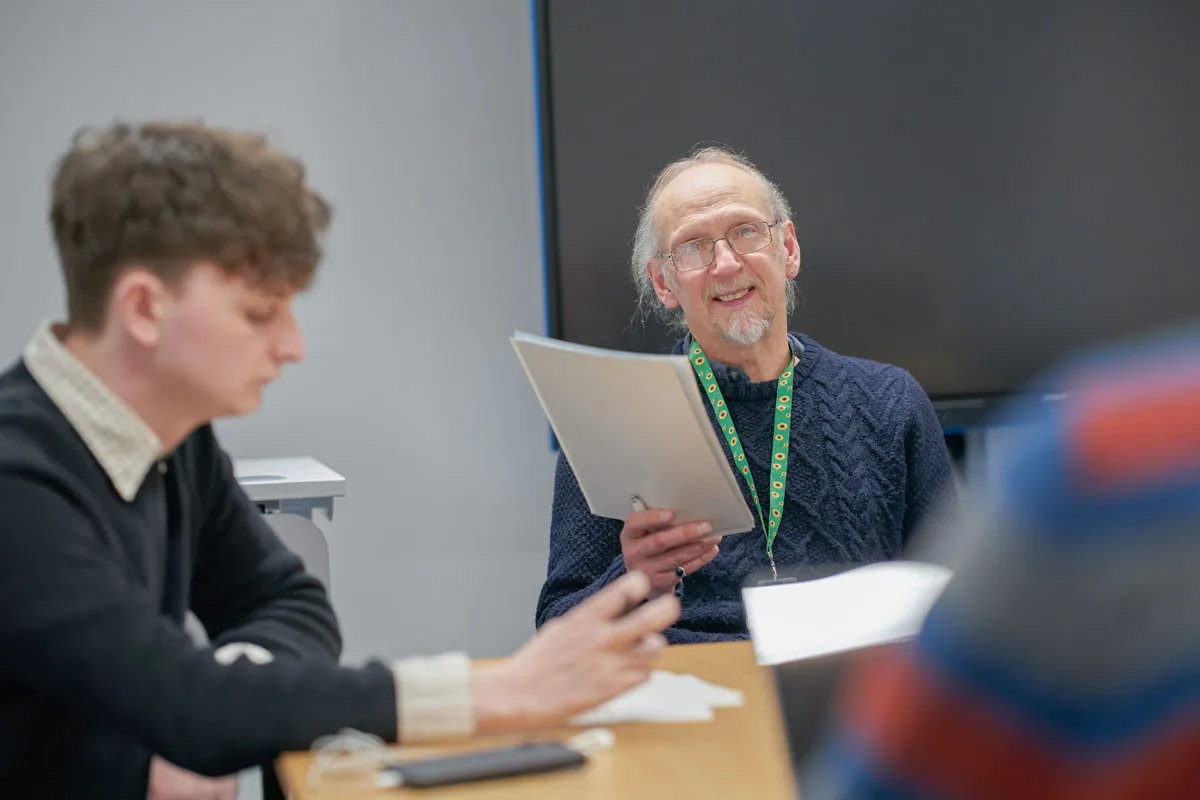 A lecturer wearing glasses and a navy jumper smiles while holding a folder in a classroom. A student in the foreground is blurred, focusing on their work, and another person’s arm is partially visible. A large screen is in the background.