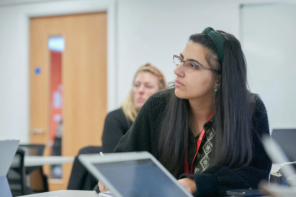 A person with long dark hair, wearing a green headband and black sweater, sits at a table in a classroom setting. They’re writing in a notepad beside a tablet or laptop. Behind them is another person with blonde hair, and the room features a wooden door with a window and a whiteboard on the wall.