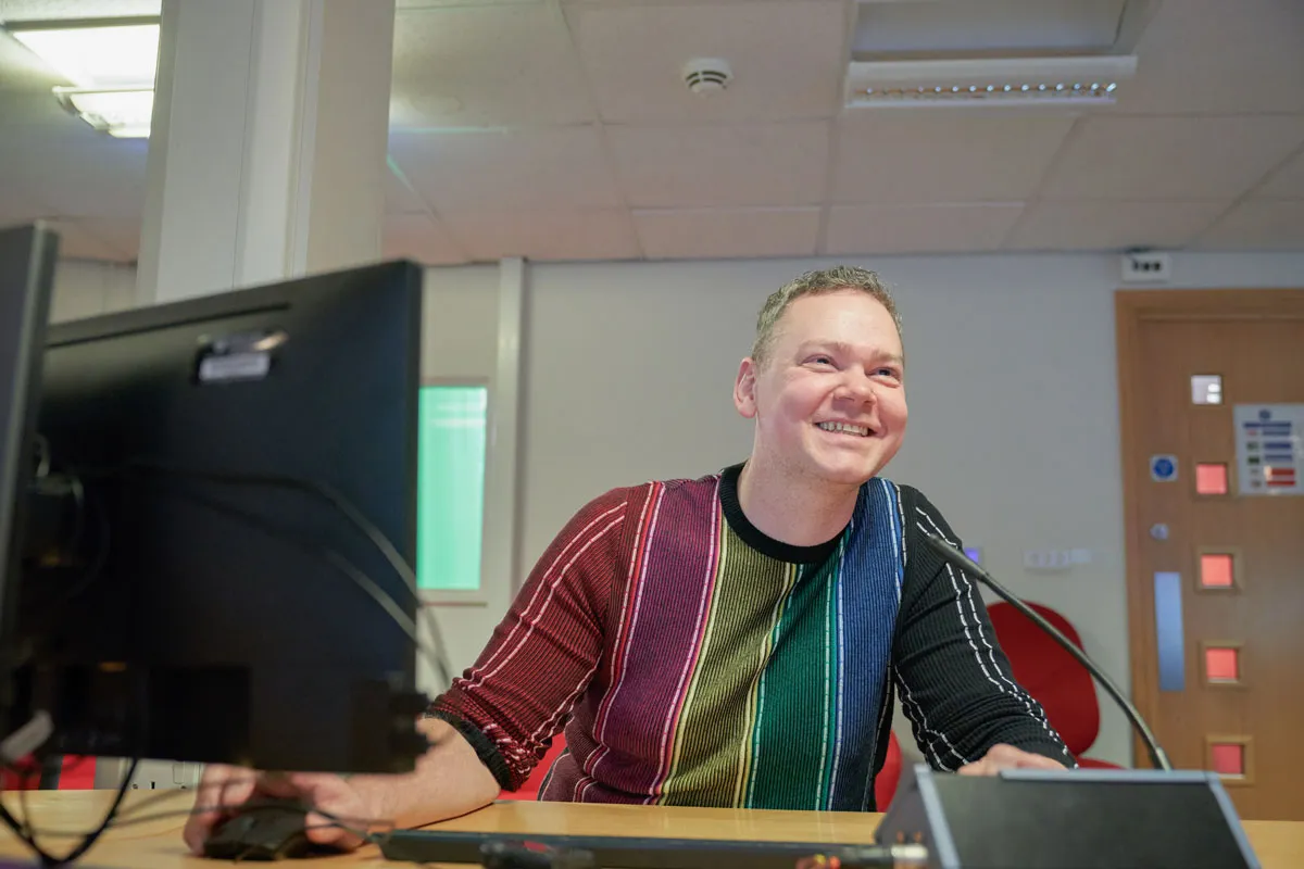 A smiling person sits at a desk in a computer lab, using a desktop computer and microphone. They are wearing a colourful, striped jumper and looking towards the screen. The background shows an office-style room with a closed door and ceiling lights.