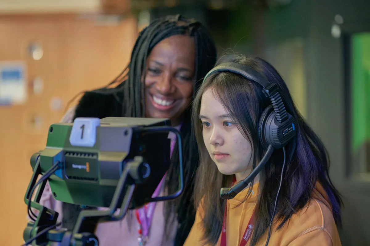 A student wearing a headset operates broadcast equipment in a media studio, focusing intently on the screen. Behind her, another student smiles while looking at the same screen. Both wear red lanyards, and the background shows a blurred studio setting.