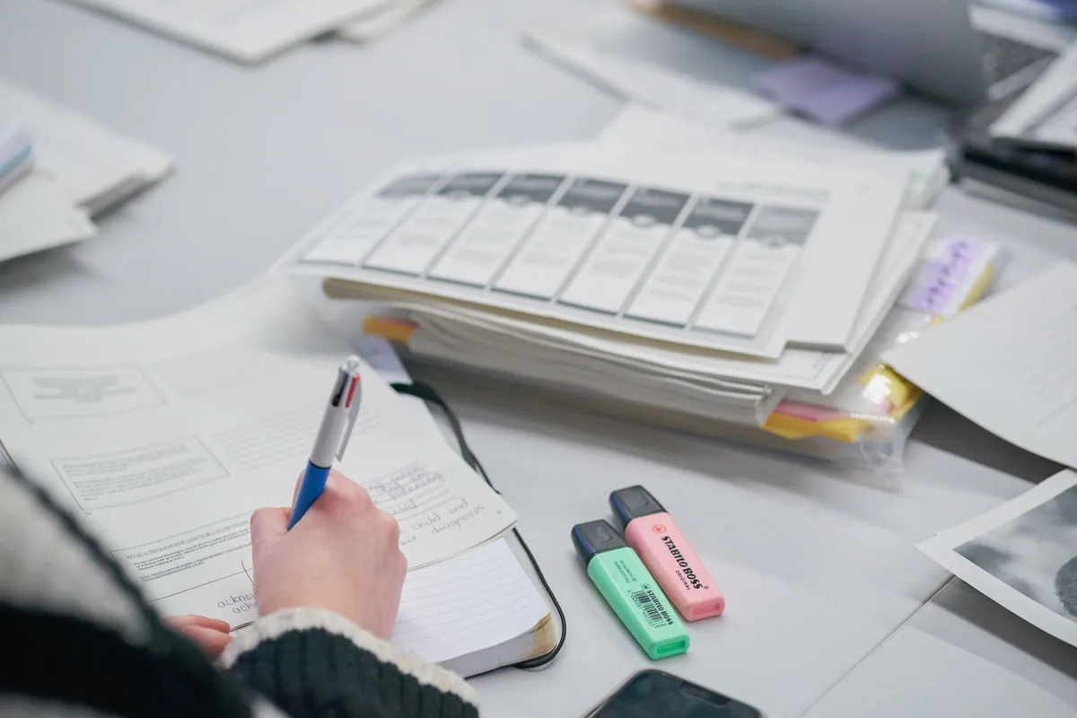 Close-up of a person’s hand writing in a notebook with a blue and white pen, surrounded by study materials including printed documents, two highlighters, and a smartphone on a desk.