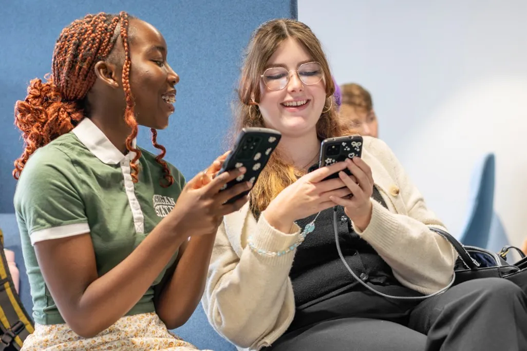 Students in Leeds City Campus Social Space laughing while looking at their phones.