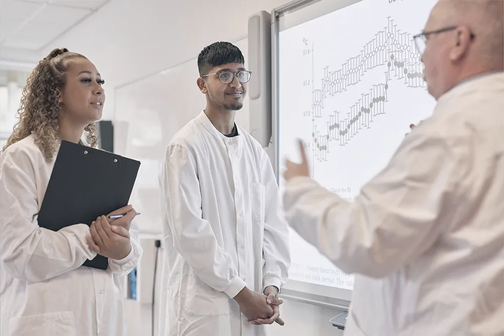 Two students in lab coats speaking to a Biomedical Science lecturer.