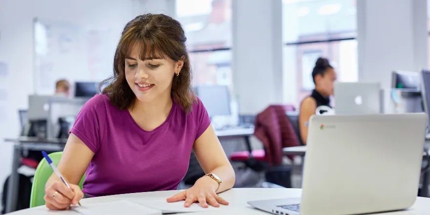 Brunette woman in purple top writing notes on paper with laptop.