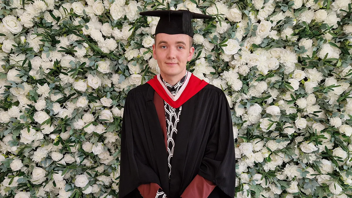 A student in graduation cap and gown smiling in front of a flower wall..