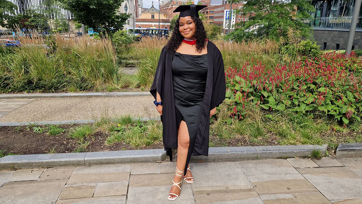 A student in graduation cap and gown smiling in a green space in Leeds..