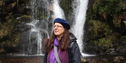 Adult female wearing purple hat stands in front of waterfall