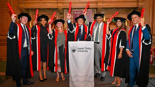 A group of people in graduation caps and gowns, holding up graduation scrolls behind a lectern covered with a white Leeds Trinity University branded cloth.