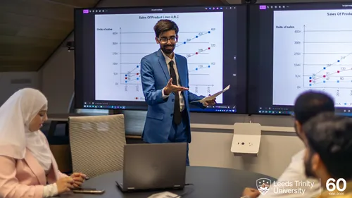 A business lecturer presenting in front of a screen. A white logo marking the 60th anniversary of Leeds Trinity University is in the bottom right corner.