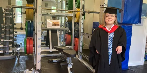 A student wearing a graduation cap and gown stands in a gym