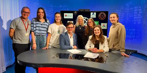 Group of male and female students and tutors stand behind red desk in front of television screen
