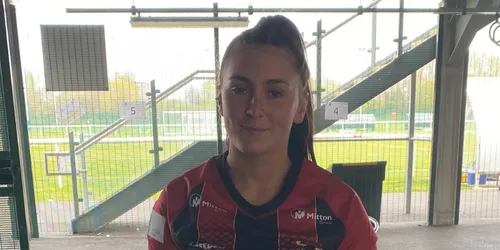 Female with brown hair tied back stands in rugby shirt in front of window