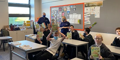 Children in classroom sat at desks holding science books with adult female and adult male stood at back