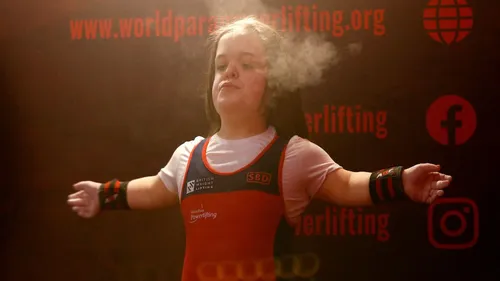 A woman with dark hair stands with her arms outstretched in front of a dark wall