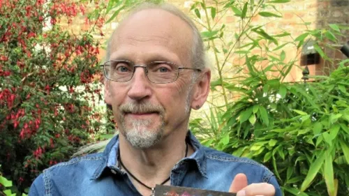 A man wearing glasses and blue shirt smiles while stood in front of green bushes