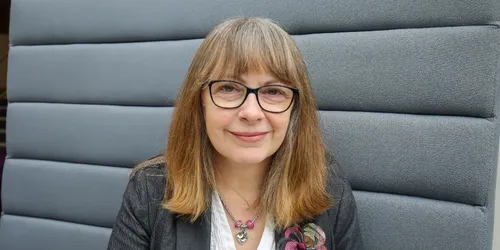 A headshot of a woman with brown hair and glasses, sitting on a grey sofa
