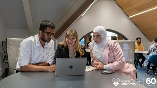 Three students working on a laptop at the Leeds Trinity University City Campus. A white logo marking the 60th anniversary of Leeds Trinity University is in the bottom right corner.