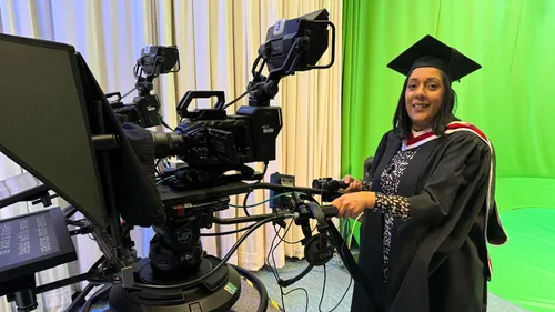 A picture of Journalism graduate Raj Kaur operating a TV camera while wearing a Graduation cap and gown