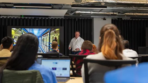 A male lecturer standing in front of a group of students in a lecture theatre.