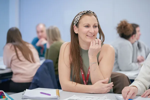 A person with long hair sits at a table in a classroom or study environment, wearing a sleeveless top and a red lanyard. They hold a pen and appear to be thinking or writing. On the table are notebooks, papers, and a purple pen. Other people are seated and working in the background.