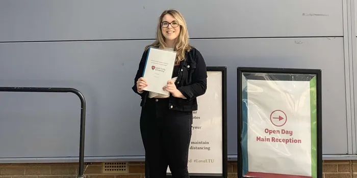 Blonde girl with black glasses in black denim jacket and jeans holds dissertation on white paper