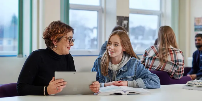 A woman with short hair and glasses holds a laptop as she teaches a girl with long brown hair sat next to her