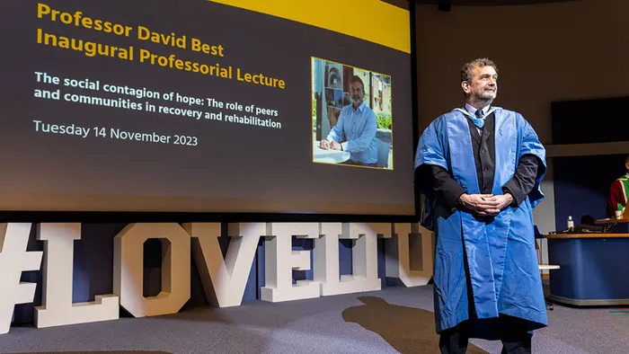 An academic in a blue gown standing in front of a screen with the title Professor David Best Inaugural Professorial Lecture written in yellow letters.
