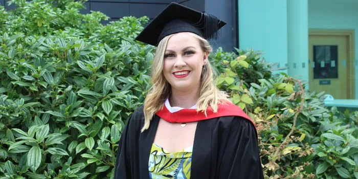 Female student stands in front of bush wearing black graduation cap and gown