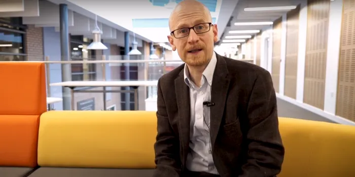 Male wearing glasses, blazer and shirt sits on yellow sofa