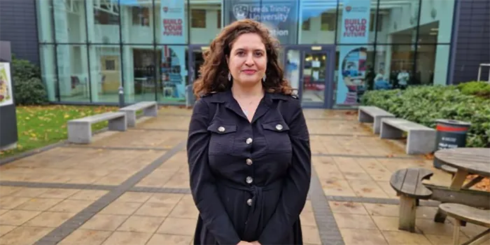 A woman with dark brown curly hair and wearing a black dress is smiling outside Leeds Trinity University.