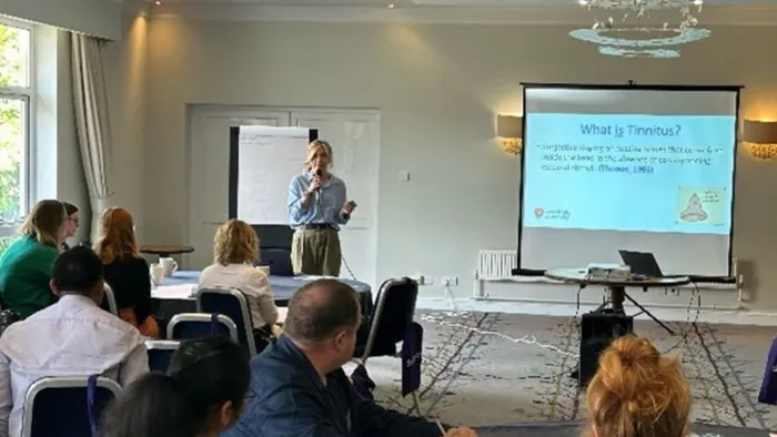 A woman with blonde hair and a blue shirt stands at the front of a room while presenting to an audience
