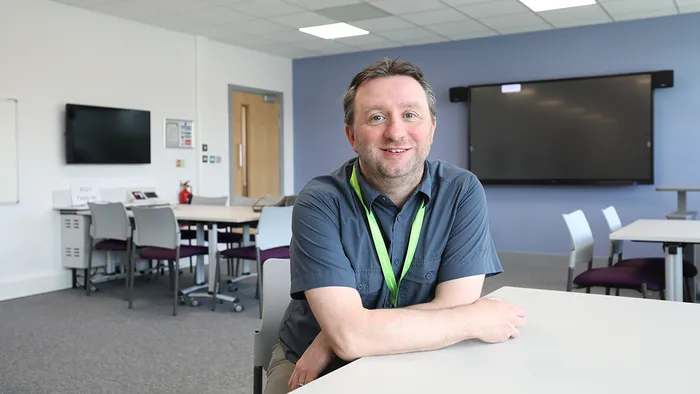 A lecturer sitting behind a desk and smiling.