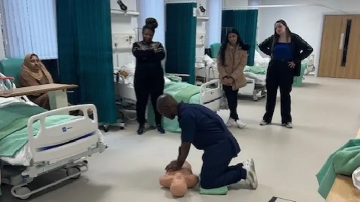 A man on his knees demonstrates chest compressions on a dummy as four students watch