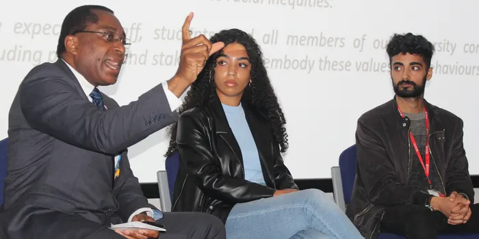 Male sits in front of screen speaking with a female and male looking at him also sat down next to him