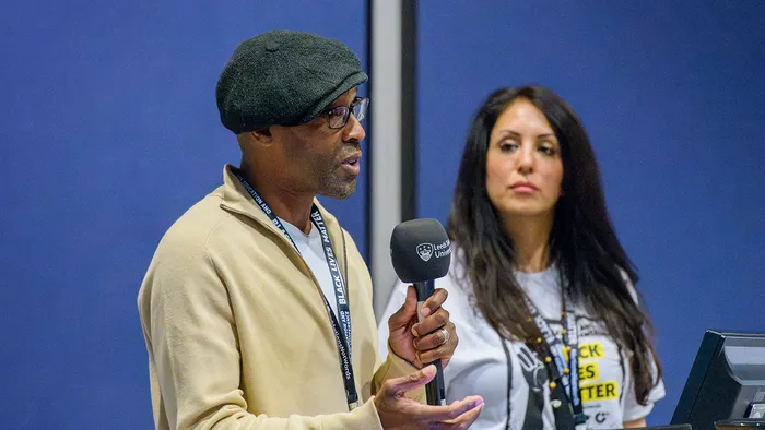 A male lecturer giving a talk and a female lecturer listening in the background.